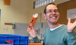 Ein Mitarbeiter der Caritas Werkstatt für Menschen mit Behinderung München hat eine leere Tabasco-Verpackung in der Hand und lacht in die Kamera | © Caritas Werkstatt für Menschen mit Behinderung München