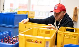 Ein Mitarbeiter mit FC-Bayern München Cap steht hinter mehreren Kisten | © Caritas Werkstatt für Menschen mit Behinderung München