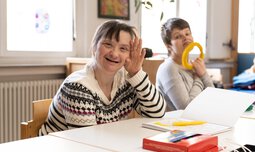 Zwei Frauen mit Behinderung sitzen vor Mal-Utensilien und schauen in die Kamera | © Caritas Werkstatt für Menschen mit Behinderung München