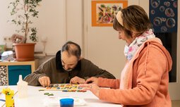Ein älterer Mann versucht Buchstaben aus Holz in eine dafür vorgesehene Einkerbung zu legen und eine Frau hilft ihm dabei | © Caritas Werkstatt für Menschen mit Behinderung München