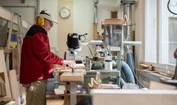Ein Mitarbeiter der Caritas Werkstatt für Menschen mit Behinderung München steht mit Ohrenschutz an einer Maschine in der Schreinerei und bearbeitet ein Holzprodukt | © Caritas Werkstatt für Menschen mit Behinderung München