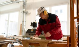 Ein Mitarbeiter der Caritas Werkstatt für Menschen mit Behinderung München steht mit Ohrenschutz an einer Maschine in der Schreinerei und bearbeitet ein Holzprodukt | © Caritas Werkstatt für Menschen mit Behinderung München