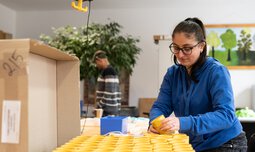 Eine Mitarbeiterin der Caritas Werkstatt für Menschen mit Behinderung München stellt fertige Produkte auf einem Tisch ab | © Caritas Werkstatt für Menschen mit Behinderung München